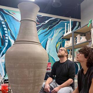 Two potters sitting and looking up at a giant unglazed vase with a flared neck beside a blue mural at The Clay Hole in Draper, Utah