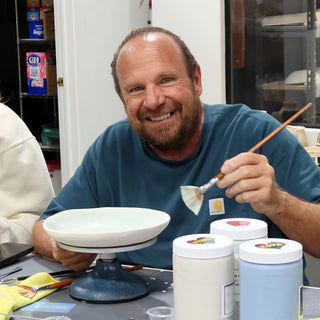 A potter grinning with a brush in hand beside a wide white plate on a turntable and jars of glaze at The Clay Hole in Draper, Utah