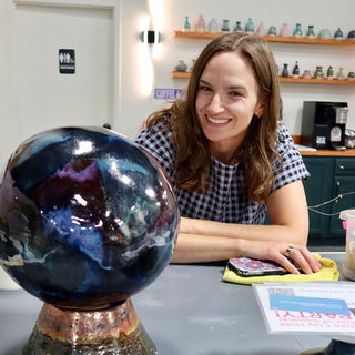A smiling potter leans on a table beside a large glossy sphere glazed in deep blue, purple and black swirls at The Clay Hole in Draper, Utah
