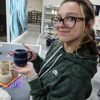 A potter in glasses and a green camouflage hoodie holding up a glossy deep blue mug beside unglazed cups at The Clay Hole in Draper, Utah