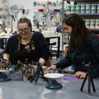 Two potters smiling at a table displaying several glossy glazed pieces in purple, brown and green with phone tripods nearby at The Clay Hole in Draper, Utah