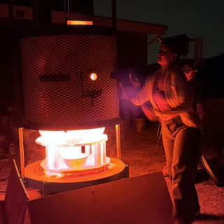 A person in a headlamp stands beside a tall mesh kiln glowing orange around a pale bowl on a stand at The Clay Hole in Draper, Utah