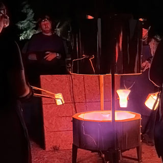 Tongs hold glowing orange pieces near a red lit kiln while a group of potters watches in the dark at The Clay Hole in Draper, Utah