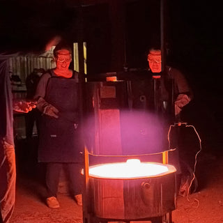 Two potters in safety goggles stand behind a kiln with a bright glowing orange opening as their faces light up at The Clay Hole in Draper, Utah