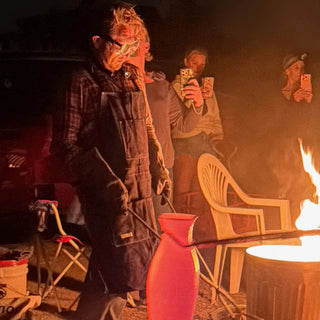 A potter in goggles and a denim apron holds tongs beside a glowing orange vase while onlookers film with phones at The Clay Hole in Draper, Utah