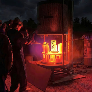 At night a person in dark clothing reaches long tongs toward glowing orange pots inside an open outdoor kiln, with The Clay Hole, a pottery studio in Draper, Utah