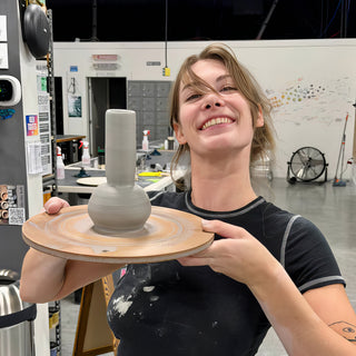 A smiling potter holds out a board with a gray vase that has a round base and tall narrow neck at The Clay Hole in Draper, Utah