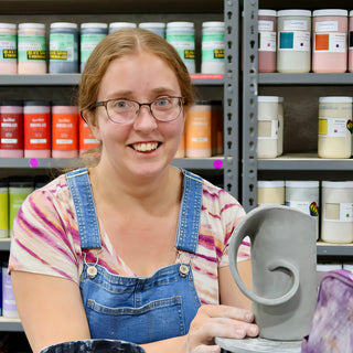 A smiling potter in denim overalls beside a gray clay vessel with a curled spiral handle at The Clay Hole in Draper, Utah