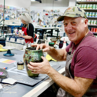 A smiling potter in a camouflage cap holding a glossy green vase with dark patches and thin light lines at The Clay Hole in Draper, Utah
