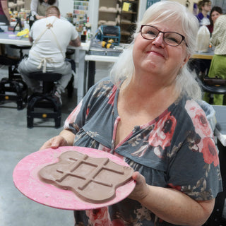 A smiling potter holding a pink board with a flat clay slab pressed with a grid of raised lines at The Clay Hole in Draper, Utah