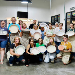A large class holding up shallow plates with pale painted designs, some kneeling and some standing at The Clay Hole in Draper, Utah