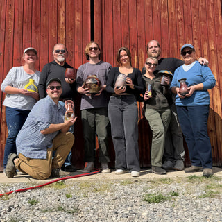 A group of members pose outdoors against a red wooden wall, each holding a small finished vase or vessel, with The Clay Hole, a pottery studio in Draper, Utah
