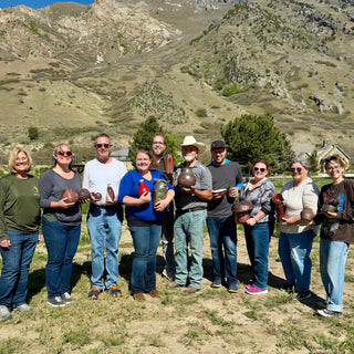A group of members line up on dry grass in front of a mountain hillside, each holding small finished pieces at The Clay Hole in Draper, Utah