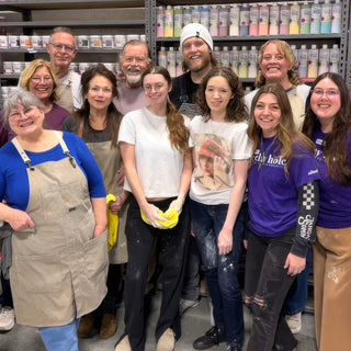A large group of members in aprons standing close together and smiling in front of shelves of glaze bottles at The Clay Hole in Draper, Utah