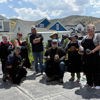 A group of potters in black aprons, goggles and heavy gloves posing outdoors, holding small dark pottery pieces, with The Clay Hole, a pottery studio in Draper, Utah