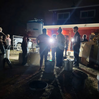A group of people stand in bright light near a red barn at night, with metal cans on the ground at The Clay Hole in Draper, Utah