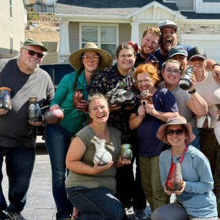 A group of members poses outdoors in front of a house, many holding small dark and copper colored vessels, with The Clay Hole, a pottery studio in Draper, Utah
