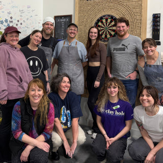 A group of potters standing and kneeling in two rows and smiling in front of a dartboard at The Clay Hole in Draper, Utah