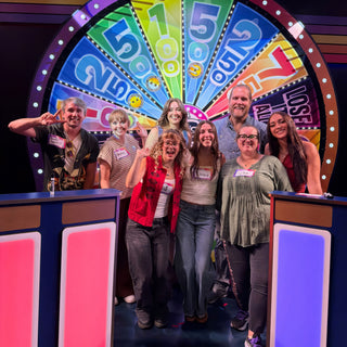 A group of smiling people with name tags pose in front of a colorful prize wheel on a stage set, with The Clay Hole, a pottery studio in Draper, Utah