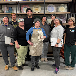 A group poses in front of shelves of plates, one in an apron holding a small white plush dog at The Clay Hole in Draper, Utah