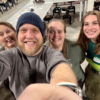 A group of four smiling potters crowd into a selfie in front of long worktables at The Clay Hole in Draper, Utah