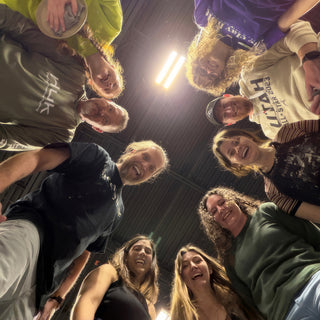 A group of smiling potters leaning in around the camera in a low-angle shot looking up toward the ceiling lights at The Clay Hole in Draper, Utah