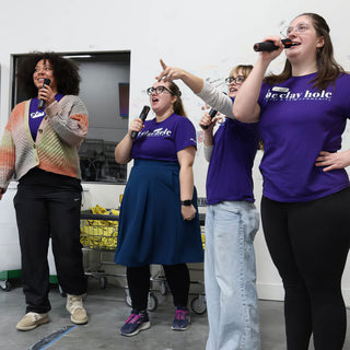 A group of four sings into microphones, three in purple t-shirts, in front of a white wall at The Clay Hole in Draper, Utah