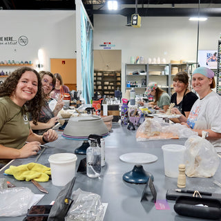 Several smiling people sit along a long gray table in an open studio, with turntables and tools set out at The Clay Hole in Draper, Utah