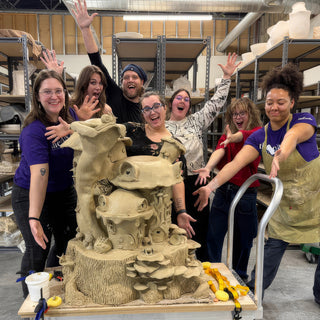 A group of seven with arms out toward a tall sculpted clay piece of mushroom houses on a stump at The Clay Hole in Draper, Utah