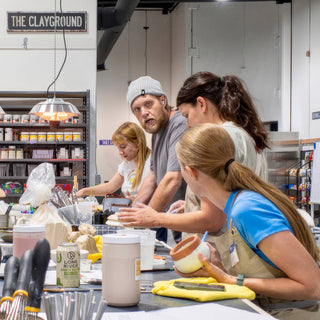 A group of potters work at a shared table while one holds a small orange cup with a white inside at The Clay Hole in Draper, Utah