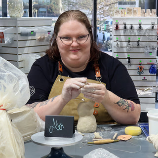 A potter in a tan apron working small clay pieces in both hands beside a bulb-shaped clay form at The Clay Hole in Draper, Utah