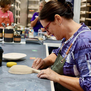 A potter in a green apron presses a shallow clay dish with a long flat handle on a table at The Clay Hole in Draper, Utah