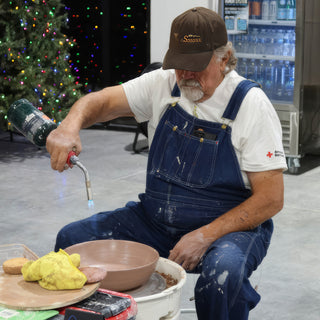 A potter in overalls and a cap holds a handheld torch above a brown bowl on the pottery wheel at The Clay Hole in Draper, Utah
