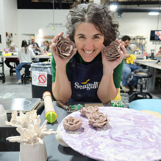 A potter in a navy apron smiling and holding up two handmade clay flowers, with more on a purple board at The Clay Hole in Draper, Utah