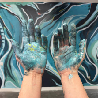 Two open hands held palms up, smeared with teal and blue color, in front of a swirled teal mural at The Clay Hole in Draper, Utah