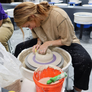 A potter leans over the wheel with both hands in a pale clay bowl beside an orange bucket at The Clay Hole in Draper, Utah