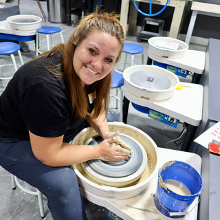 A smiling potter leans over a pottery wheel with both hands on a small lump of clay at The Clay Hole in Draper, Utah