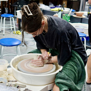 A potter with a claw hair clip leans over a wheel with both hands on a lump of clay at The Clay Hole in Draper, Utah