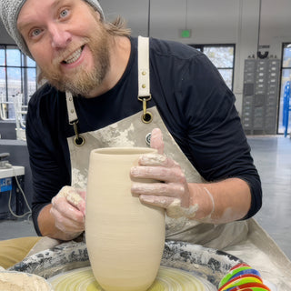 A grinning potter wraps both hands around a tall, straight-sided clay vase with fine ridges on a pottery wheel at The Clay Hole in Draper, Utah