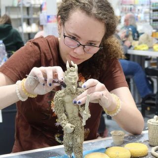 A potter with glasses detailing a small standing clay figure with horns and a rough textured body at The Clay Hole in Draper, Utah