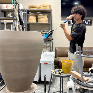 A potter aiming a long blue torch at a huge unglazed gray-brown vase standing in the studio at The Clay Hole in Draper, Utah