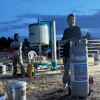 Two people stand outdoors at dusk beside a tall gas cylinder and a round kiln with a mesh top, with The Clay Hole, a pottery studio in Draper, Utah