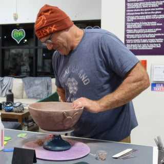 A potter in an orange beanie works on a large reddish brown bowl with a raised design on a turntable at The Clay Hole in Draper, Utah