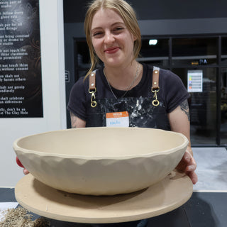 A smiling potter holds a large wide unglazed bowl with faceted sides resting on a round board at The Clay Hole in Draper, Utah