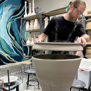 A potter holds a wide flat gray clay form over a large open unfired vessel, with a blue mural behind at The Clay Hole in Draper, Utah