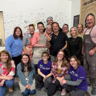 A large group of members posing in two rows in front of a wall covered in handwritten signatures and doodles at The Clay Hole in Draper, Utah