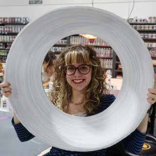 A smiling potter looks through the hole of a very large white ring-shaped piece held up with both hands at The Clay Hole in Draper, Utah