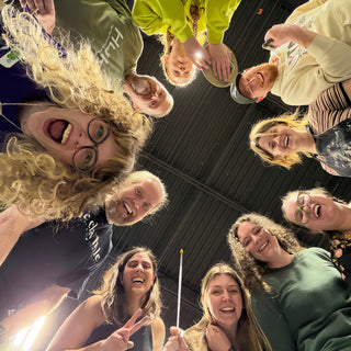 A circle of laughing potters seen from below, with one in curly hair and round glasses up front at The Clay Hole in Draper, Utah