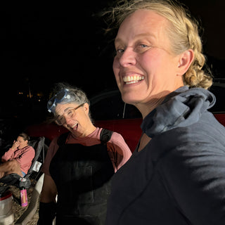 Two friends laugh toward the camera at night, one with safety goggles pushed up on their head at The Clay Hole in Draper, Utah