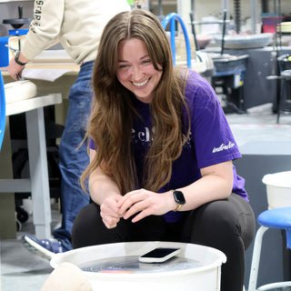 A potter in a purple t-shirt laughing with hands clasped beside a pottery wheel with a phone on it at The Clay Hole in Draper, Utah
